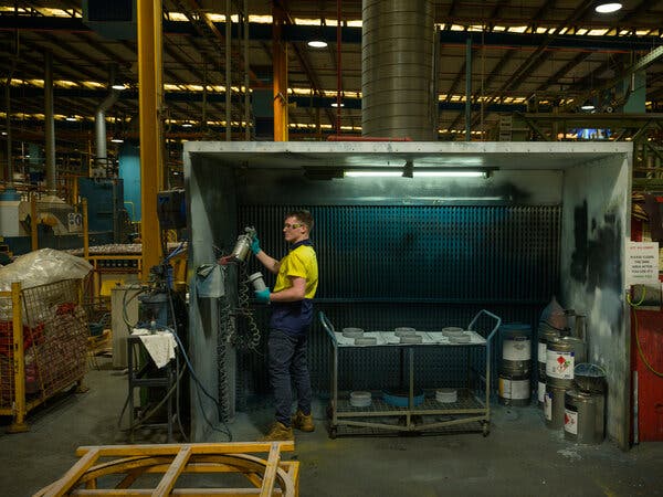 A worker in a yellow shirt holds a canister in a plant. He is inside a structure open on one side. 